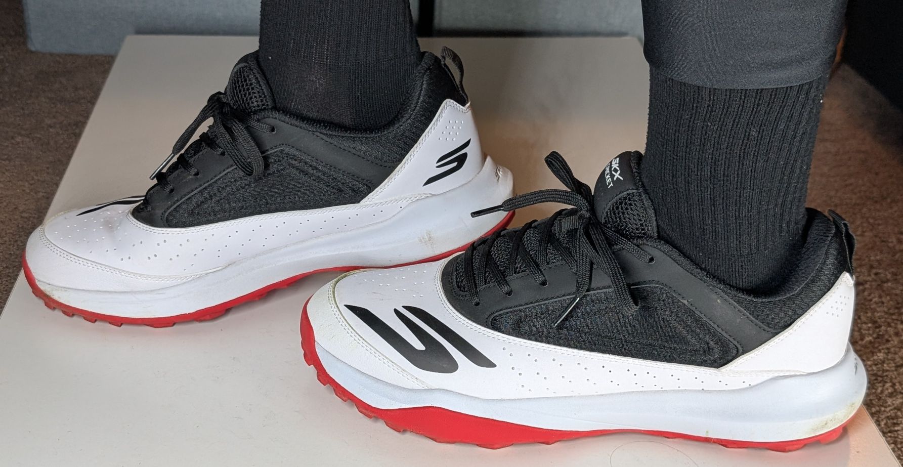 Black, white, and red Skechers Turf Cricket Shoes on feet with black socks