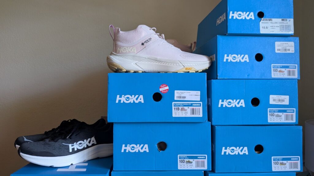 Black Hoka Solimar 2 and Pink Transport Chukka shoes on stack of blue Hoka boxes with sale stickers