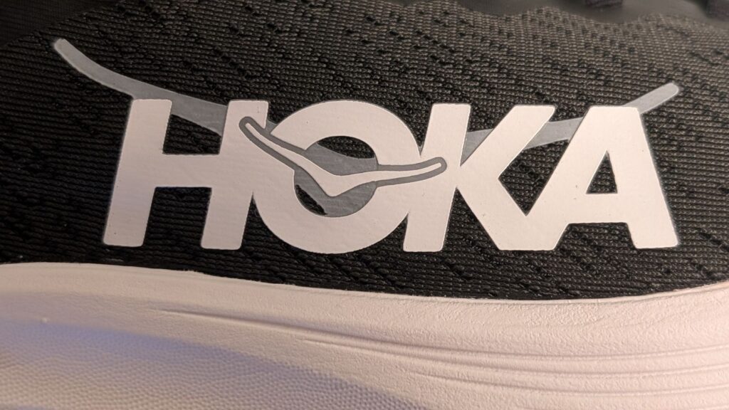 Uneven Hoka logo in white and silver on black shoe