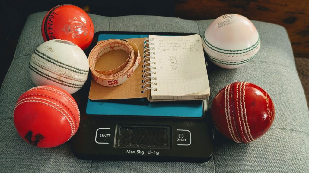Red, white, and pink hard cricket balls and synthetic cricket balls with scale and measuring tape.