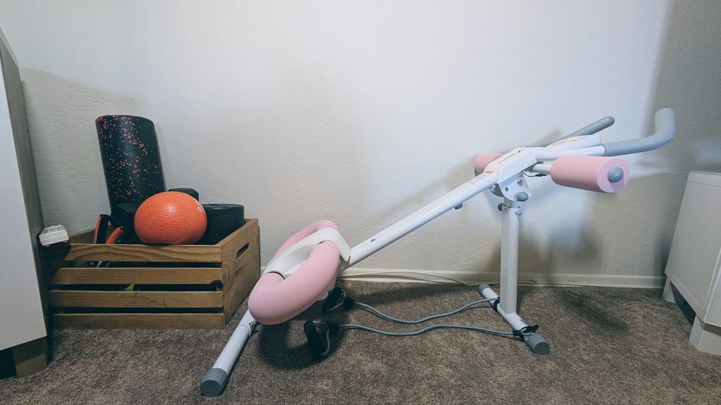 Pink and white MERACH ab trainer next to home gym foam roller and home equipment to show size